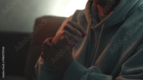 Young man in dim room suffering from hand pain, massaging wrist, clenching fist, and doing circular hand movements to relieve pain. Concept of injury, strain, recovery, muscle tension, and self treatm