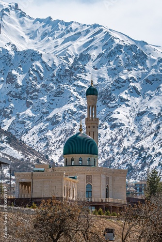 A beautiful mosque with a green dome and minaret set against a backdrop of majestic snowy mountains under a clear sky.