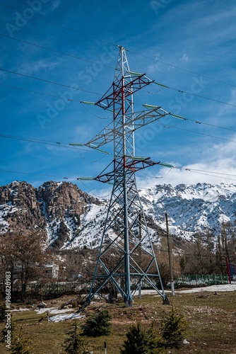 A tall power line tower stands prominently against a backdrop of snow-covered mountains and a clear blue sky, showcasing the intersection of nature and infrastructure.