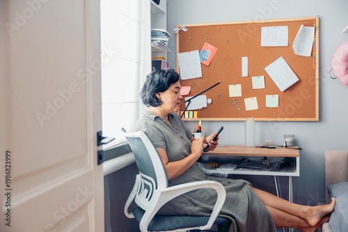 middle-aged Asian woman with short black hair sits at a desk, using a smartphone. A bulletin board with notes and papers is in the background.