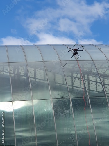 Canvas Print A tethered inspection drone hovers beside a curved glass roof of a modern building, conducting maintenance and window cleaning under a bright blue sky with scattered clouds, urban drone technology
