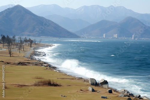 Coastal landscape showing a vast ocean with powerful waves crashing onto a rocky shore, a barren treeline, and distant mountains overlooking an urban skyline