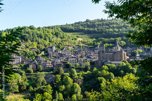 Medieval village of Conques perched on a green hillside at golden hour, panoramic view from a hill, soft evening light, France