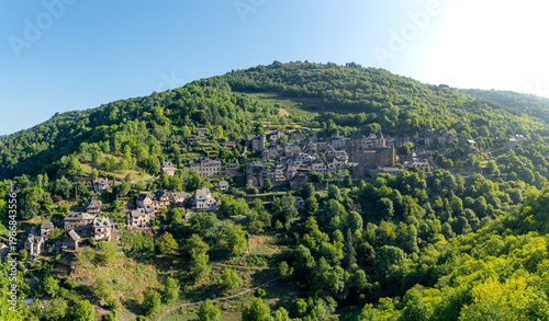 Medieval village of Conques perched on a green hillside at golden hour, panoramic view from a hill, soft evening light, France