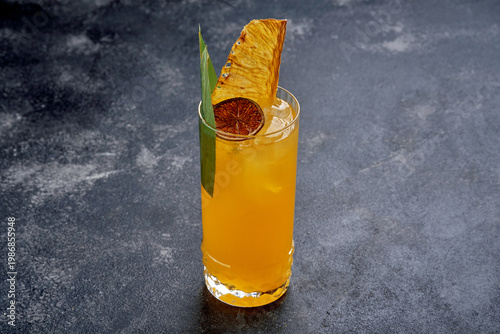 Yellow tropical fruit cocktail with ice, dried pineapple and orange garnish in a tall glass on dark background