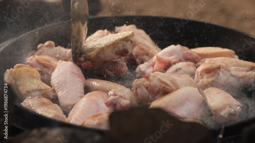 Chef stirring fresh chicken wings in a hot cast-iron skillet over an open fire while camping outdoors.