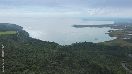 Aerial view of Lake Taupo, New Zealand, showcasing its vastness and the surrounding lush green forests and wetlands. The tranquil waters reflect the sky, creating a serene landscape.