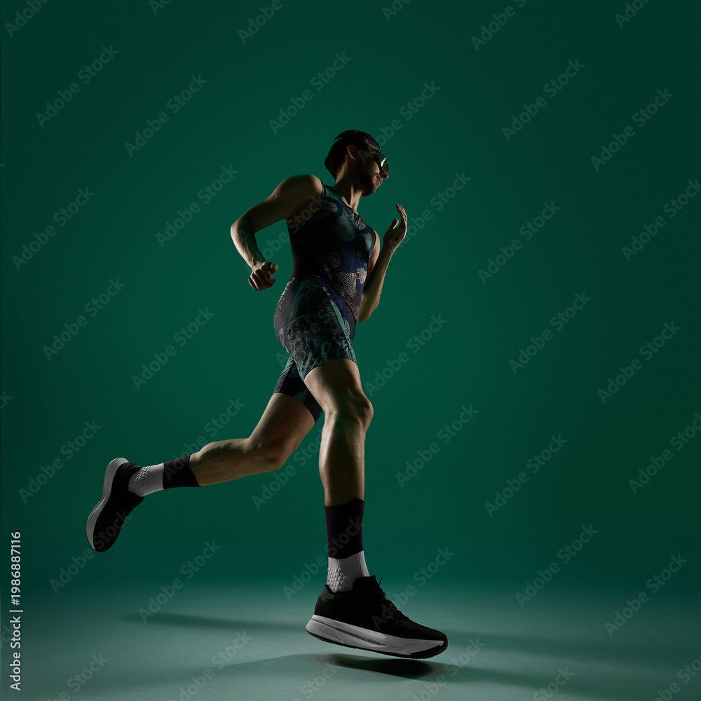 Fototapeta premium Male athlete stepping forward in running low angle motion on dark green background. Concept of fitness progress visuals, training routine promotion, active lifestyle content, sports branding material.