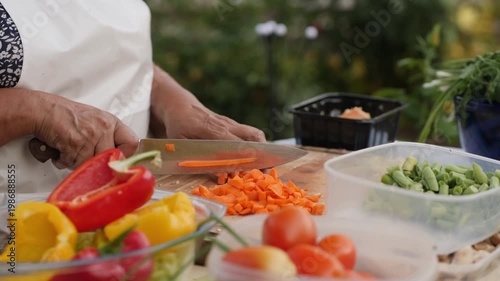 Chef slicing fresh carrots with a knife on a wooden board outdoors, preparing ingredients for a vegetable stew.