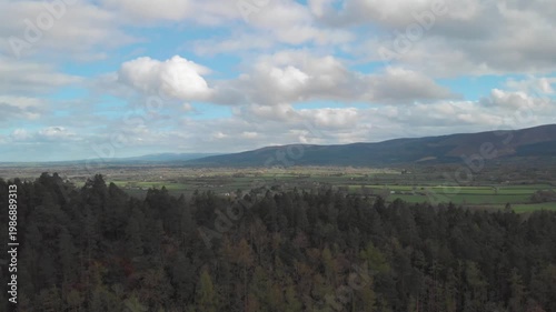 Gliding over a serene forest landscape, revealing expansive green fields and rolling mountains under a dramatic cloudy sky, inspiring peace and natural beauty.