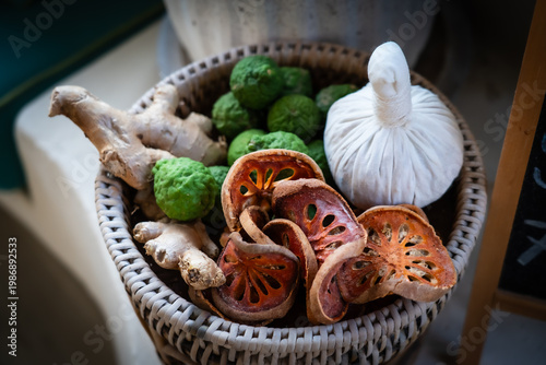 Thai herbal spa set with herbal compress ball, dried bael fruit, bergamot, and fresh ginger in a wicker basket.