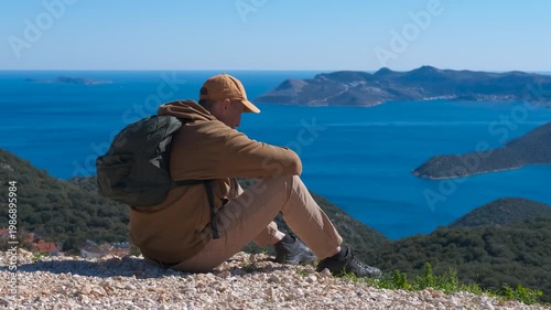 Hiker sitting on a mountain top enjoying the stunning sea view. Young male traveler with a backpack sitting on top of a mountain, contemplating the beautiful seascape with green islands