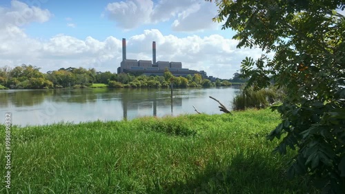 Huntly Powerstation, Huntly, Waikato, New Zealand, with tall smokestacks stands on the banks of the Waikato river, reflecting the cloudy sky.