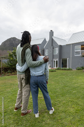 Couple embracing on grassy lawn, facing modern gray house, denim jacket and scarf