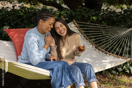 Diverse female friends sitting on hammock in garden wearing sweaters and jeans, holding glass