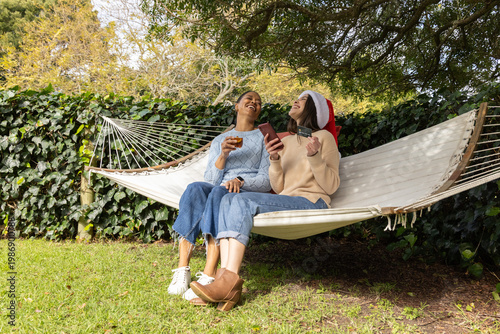 Diverse female friends sitting on hammock in garden, one wearing Santa hat, sharing phone and card
