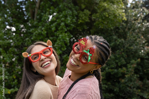 Diverse female friends leaning together laughing in garden with novelty glasses and knit sweaters
