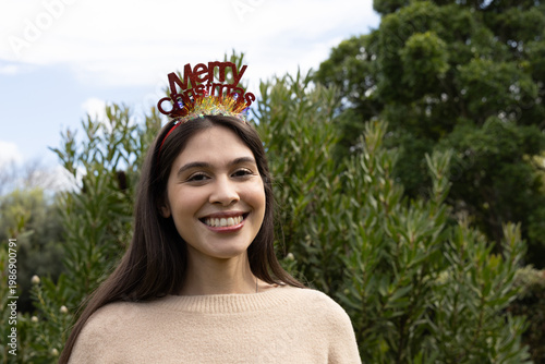 Woman smiling and wearing Merry Christmas headband while standing in park with evergreen shrubs