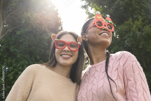 Diverse female friends standing in sunlit park, wearing reindeer and tree glasses, knit sweaters