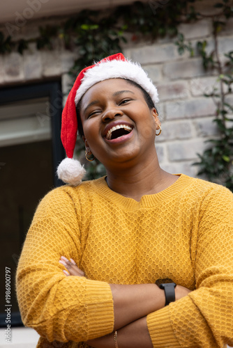 African American woman laughing wearing Santa hat yellow sweater watch and hoops by brick wall