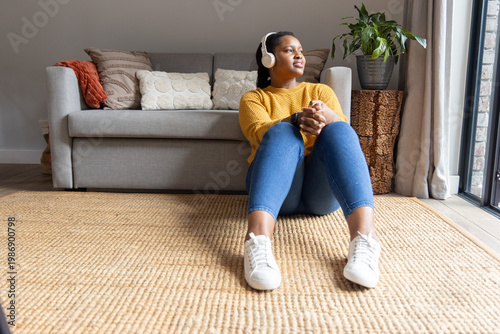 African American woman sitting on woven rug in living room wearing mustard knit headphones