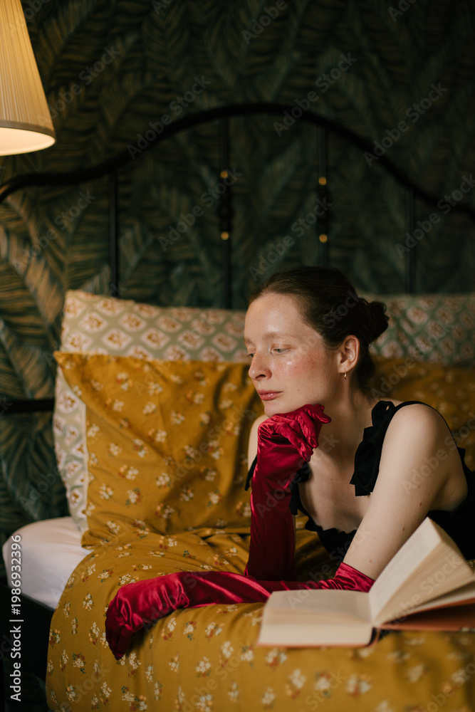 Fototapeta premium Elegant woman with frecklesin black dress and red gloves reading book in vintage interior with warm light.