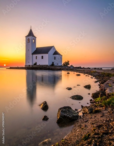 A serene shot of a white building with a steeple near tranquil water, mirroring the sunrise's vibrant hues. The rocky shoreline adds depth