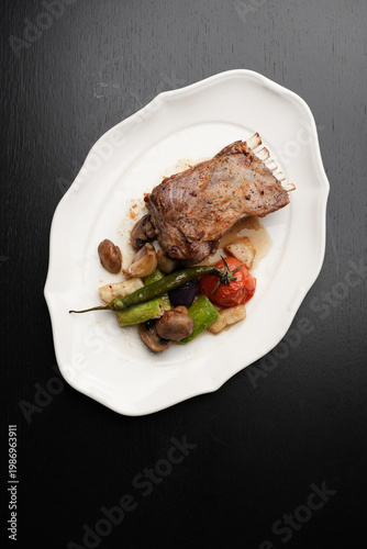 Roasted Lamb Rack with Grilled Vegetables and Chili Pepper. Baked lamb ribs served with grilled tomatoes, zucchini, mushrooms, and a whole green chili pepper. Gourmet meat dish on a white plate
