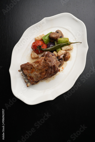 Roasted Lamb Rack with Grilled Vegetables and Chili Pepper. Baked lamb ribs served with grilled tomatoes, zucchini, mushrooms, and a whole green chili pepper. Gourmet meat dish on a white plate