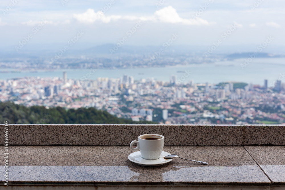 Obraz premium Hot coffee cup on balcony overlooking Penang Hill. Morning beverage with cityscape view in Malaysia. Scenic urban landscape from elevated cafe terrace.