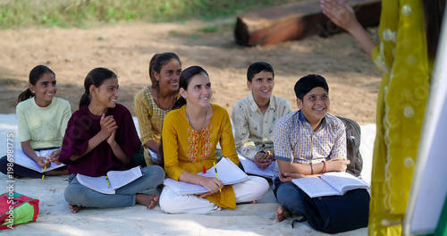 Indian adult female lady talk speak teach to villager group day under tree girl boy sitting on mat do study look board learn at open class field rural area place