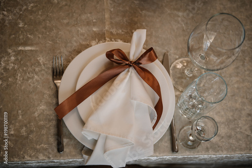 Elegant banquet table setting with stacked white ceramic plates and a folded white napkin tied with a brown satin ribbon. Placed on a textured cream velvet tablecloth with various glasses.