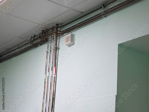 Metal pipes running along a light green wall with a ventilation grate. Industrial plumbing system inside a building. Infrastructure utility supply in a commercial office interior space.