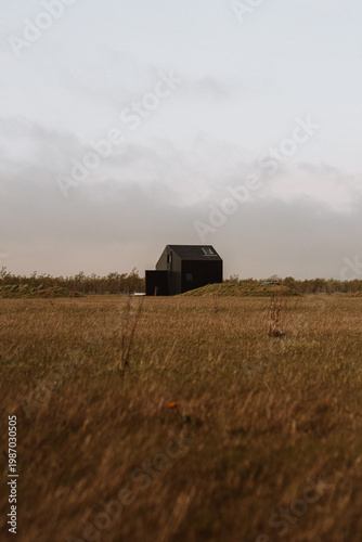 Small modern black cabin standing alone in a wide Icelandic field under a cloudy sky