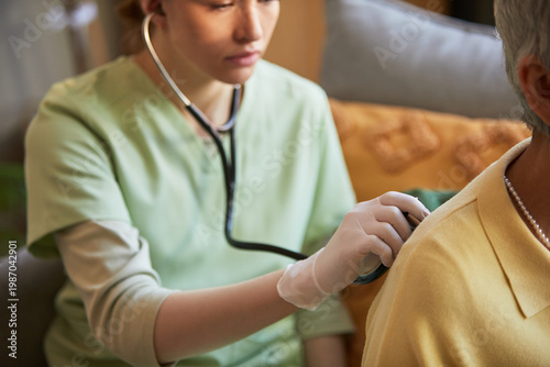 Photos Close up of young adult female caregiver checking senior woman with stethoscope