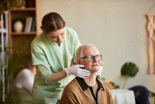Medium shot of young adult female caregiver supporting senior man during neck mobility check in nursing home