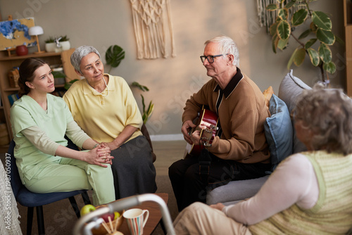 Foto Senior man playing guitar for senior women and young adult caregiver in nursing
