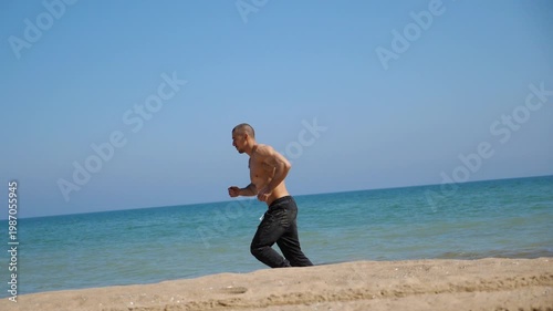 Shirtless man jogging along sandy beach, showcasing dynamic movement and energy, with gentle waves lapping at shore under clear blue sky, capturing the essence of outdoor fitness and vitality
