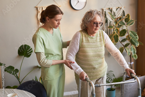 Photos Senior woman using walker while young adult female caregiver supporting mobility