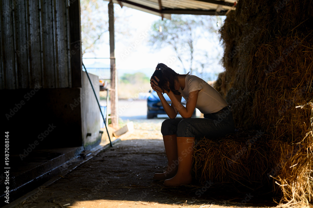 Fototapeta premium Exhausted female farm worker sitting on hay bale in barn feeling stressed and overwhelmed