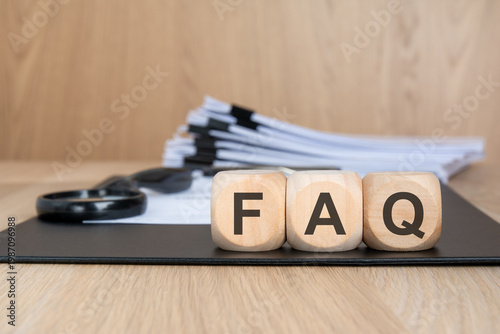 FAQ acronym on wooden blocks. Blurred background with a document stack and magnifying glass. Customer support concept.