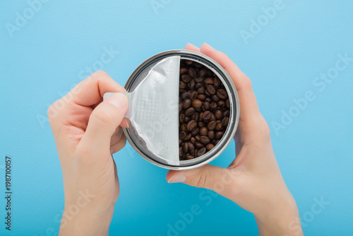 Young adult woman hands holding and opening can with fresh dark brown coffee beans on light blue table background. Pastel color. Closeup. Point of view shot. Top down view.