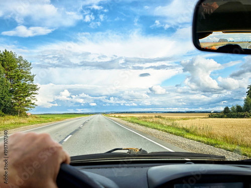 view through car windshield of natural landscape with green field, white clouds in blue sky. Hand of woman on steering wheel of car. Female traveler