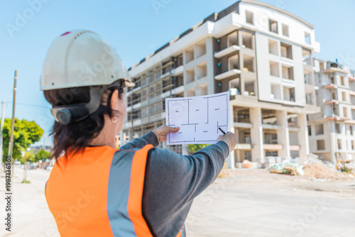 фотографія A young Hispanic woman in a hard hat and safety vest examines blueprints at a construction site with a partially built apartment building in the background