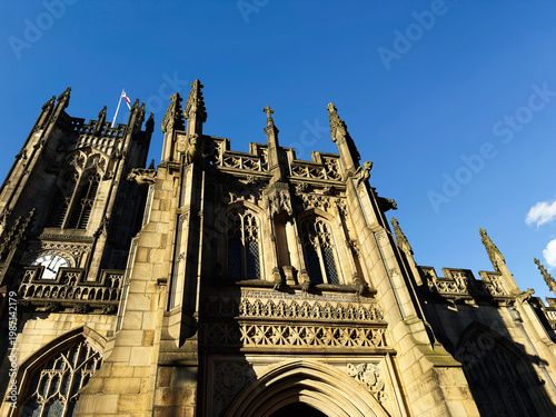 Historic Gothic cathedral facade in Manchester, UK with ornate stone architecture, carved details, towers and clock under a clear blue sky