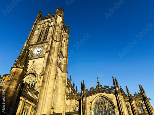 Sunlit Gothic cathedral clock tower in Manchester, UK with ornate stone architecture and stained glass windows against a clear blue sky