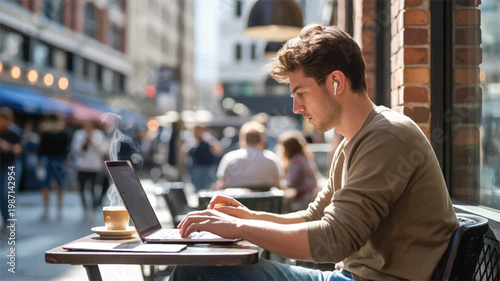 Young man working on laptop at outdoor cafe table with coffee and earbuds