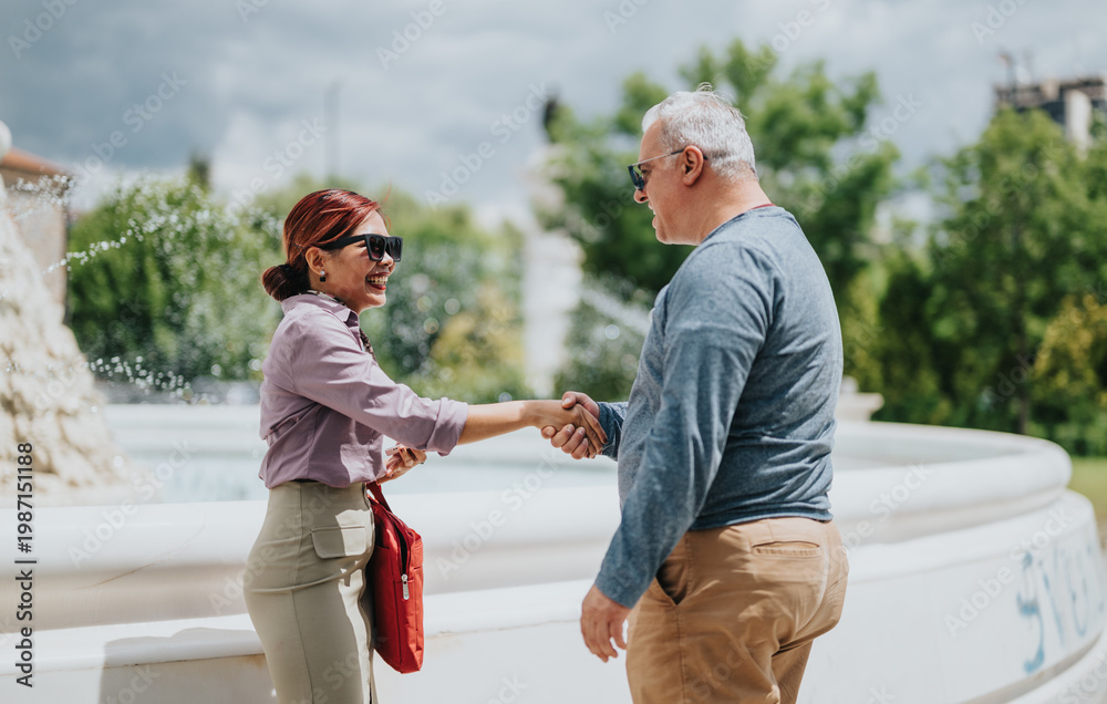 Obraz premium Two colleagues exchange a friendly business handshake and smile beside a public fountain. A casual outdoor meeting shows a professional greeting between two people.