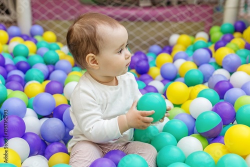 Toddler holding colorful ball while sitting in ball pit. Motor skills development.