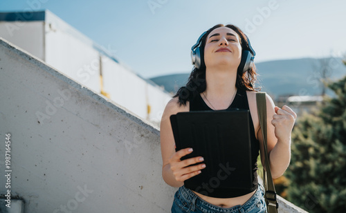 Young female student wearing headphones holds a tablet and smiles with her eyes closed. She enjoys music outdoors, raising a fist in a joyful, relaxed moment.